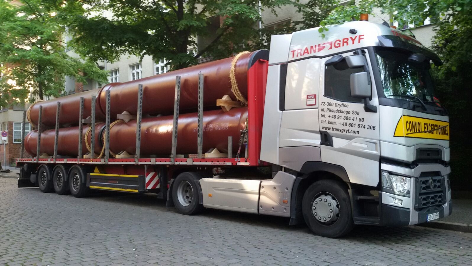Ciężarówka firmy Trans-Gryf przewożąca dwa duże, brązowe zbiorniki cylindryczne na platformie, z żółtą tablicą 'CONVOI EXCEPTIONNEL' na kabinie.