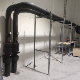 Mateusz Bartczak - Czarne, zaizolowane rury grzewcze z manometrami, biegnące wzdłuż ściany z blachy trapezowej w hali, podtrzymywane przez metalowy stelaż.