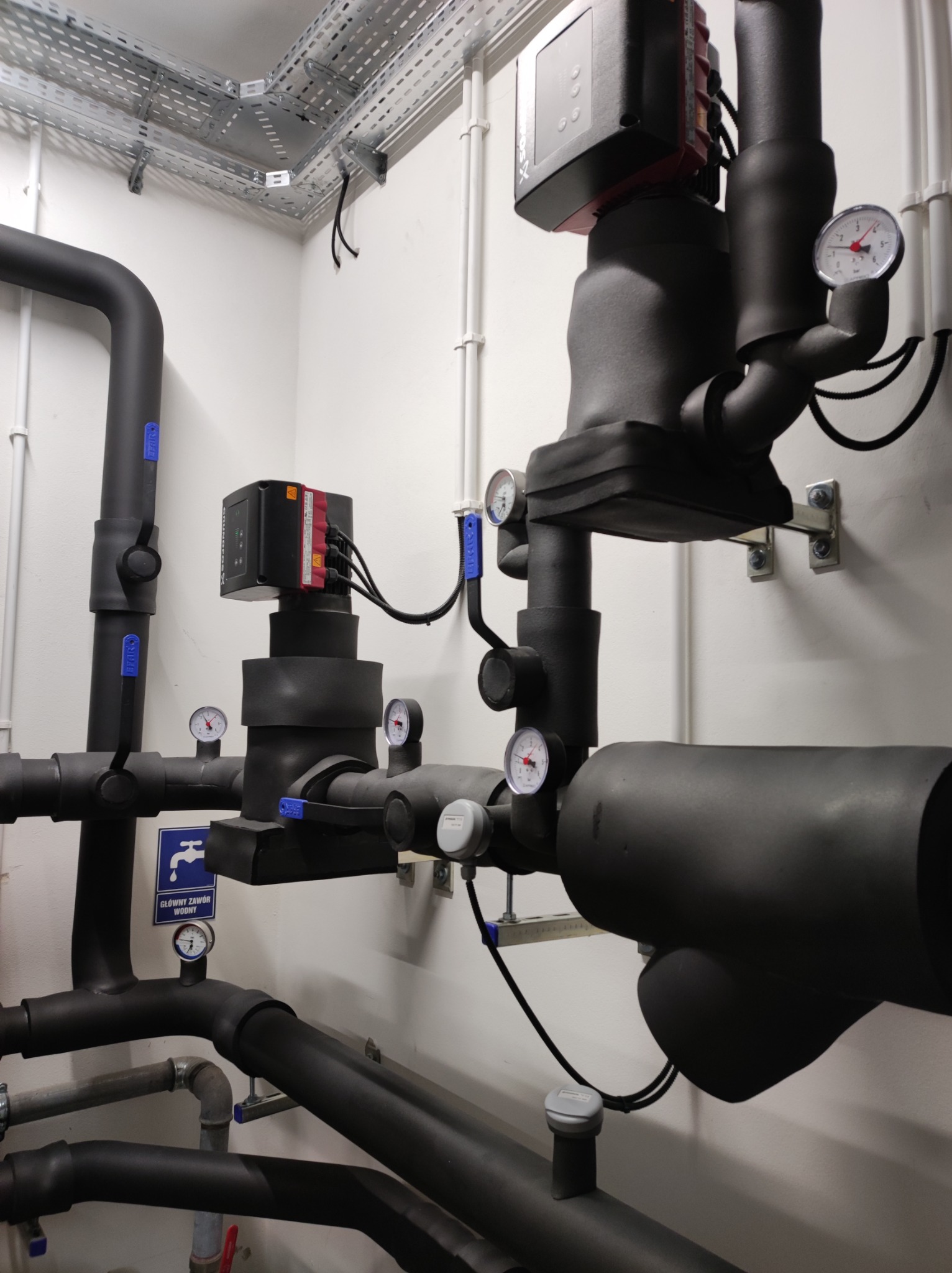 Czarno-szare rury grzewcze z izolacją termiczną, manometry i automatyka w kotłowni, widoczne elementy orurowania pod sufitem i tabliczka informacyjna.