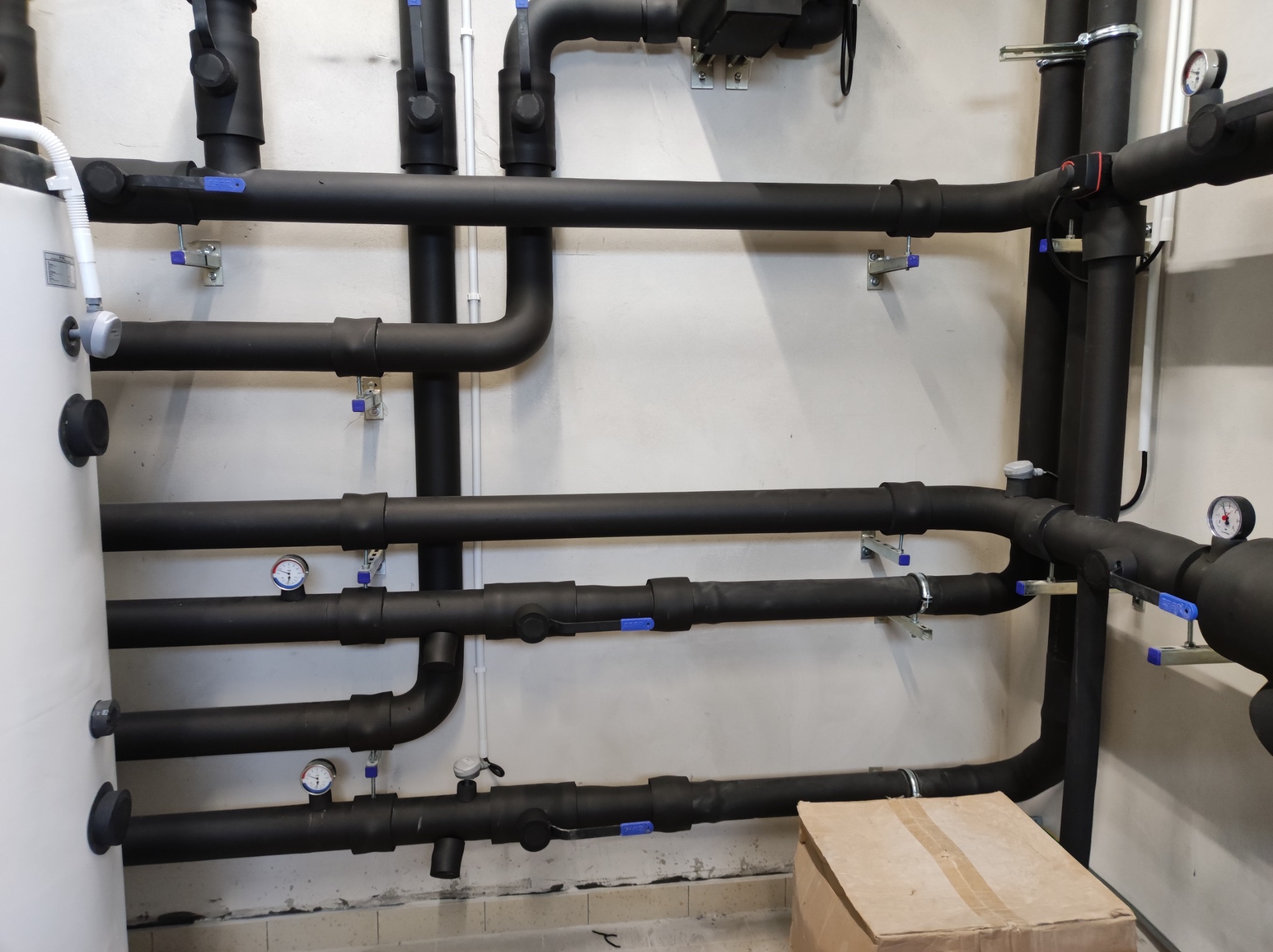Czarny system rur izolowanych termicznie z niebieskimi zaworami i manometrami zamontowany na ścianie, widoczny fragment białego zbiornika i kartonowe pudełko na podłodze.