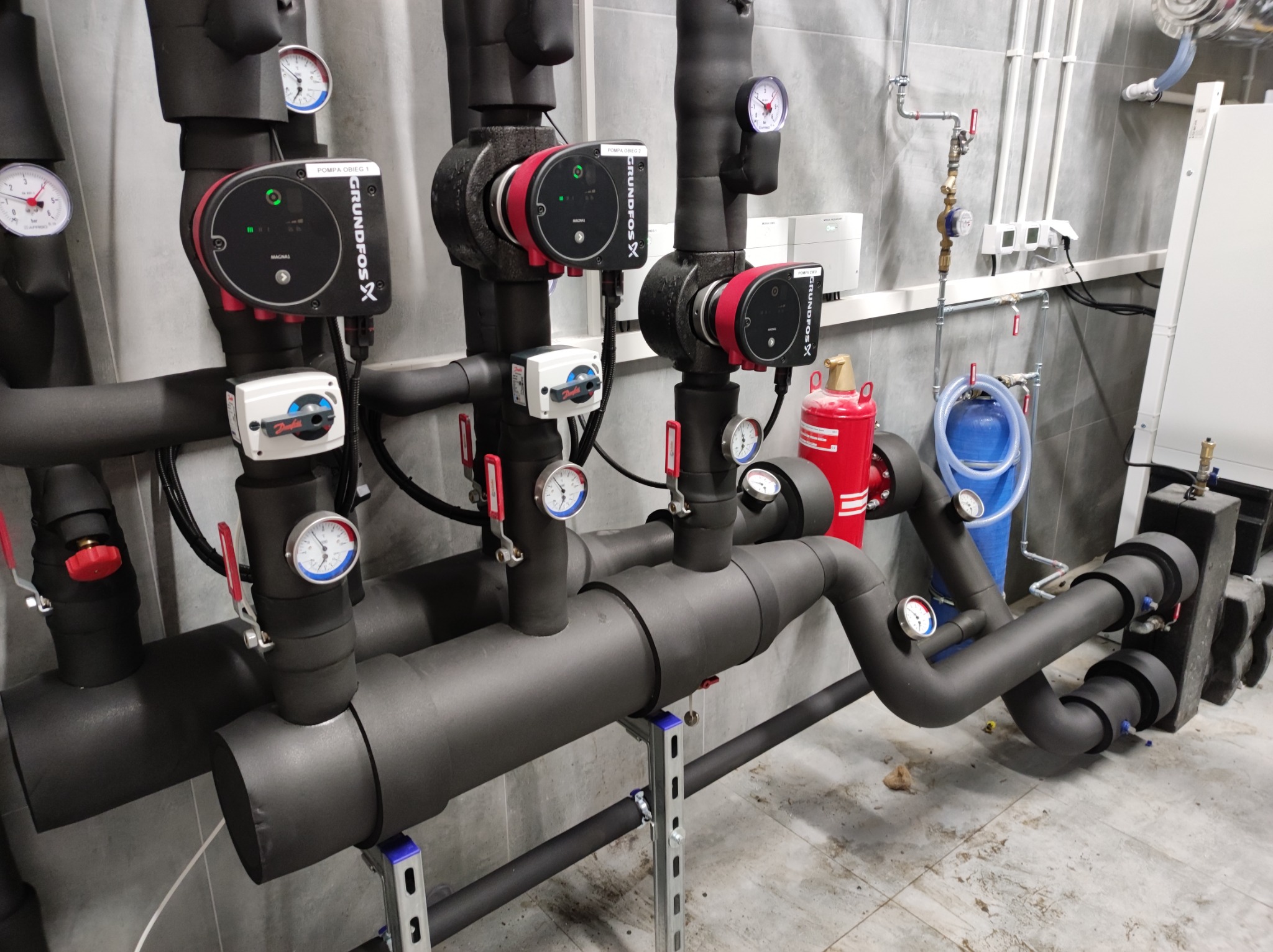 Kompleksowa instalacja grzewcza z pompami obiegowymi Grundfos, rurami w czarnej izolacji termicznej, manometrami i zaworami, zainstalowana w pomieszczeniu technicznym z szarymi płytkami.