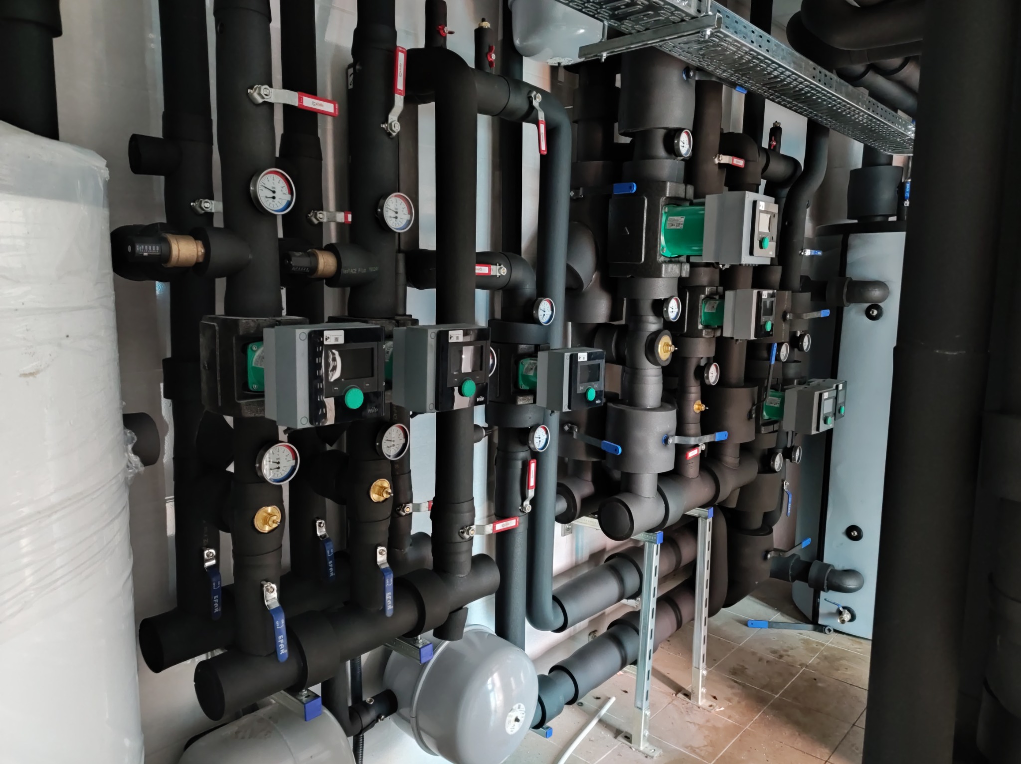Kompleksowa instalacja grzewcza z czarnych rur izolowanych termicznie, z manometrami, zaworami EFAR i szarymi pompami obiegowymi Wilo, zamontowana w kotłowni.