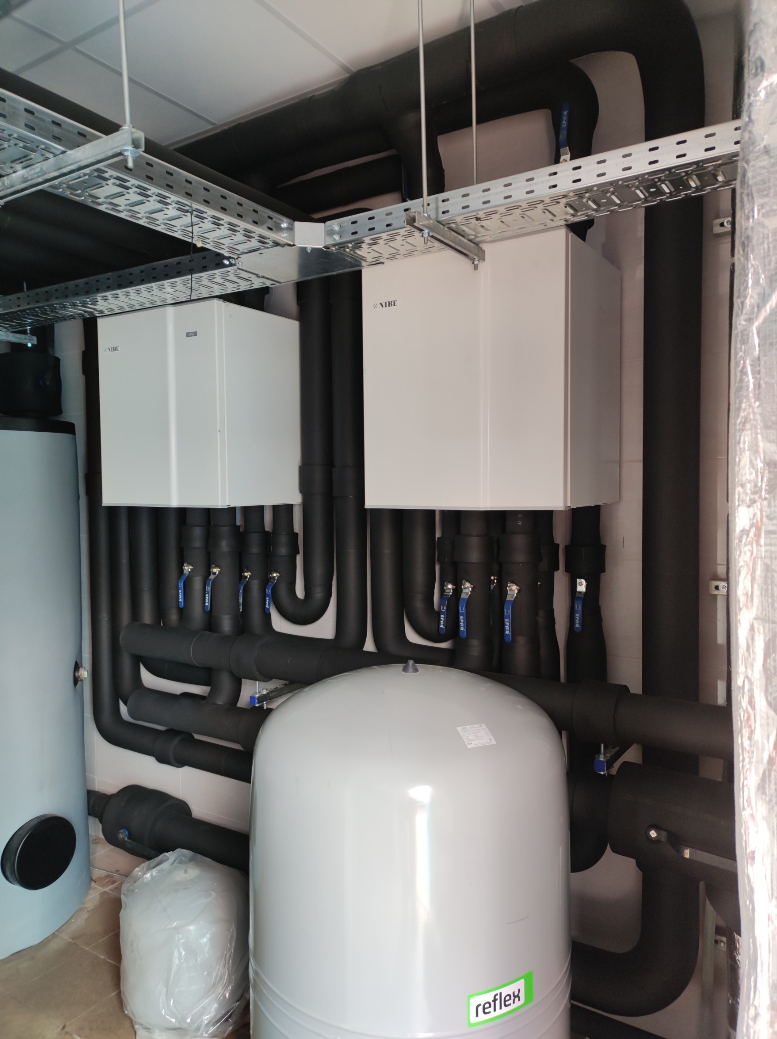 Instalacja grzewcza z dwoma białymi modułami NIBE, czarnymi rurami izolowanymi i białym zbiornikiem Reflex w pomieszczeniu technicznym z metalowymi korytami kablowymi pod sufitem.