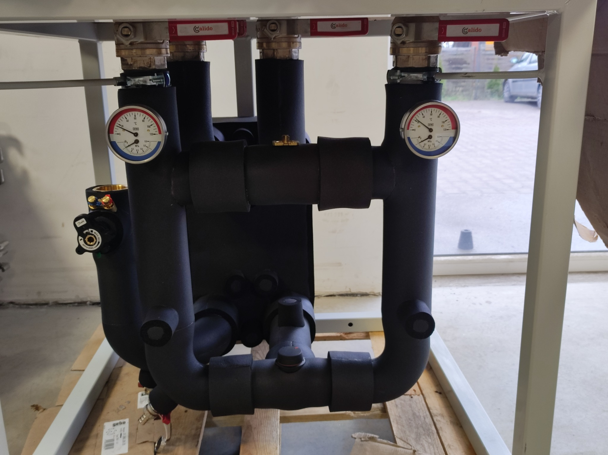 Węzeł cieplny z izolowanymi rurami, manometrami i zaworami Calido, zamontowany na drewnianej palecie w pomieszczeniu technicznym.