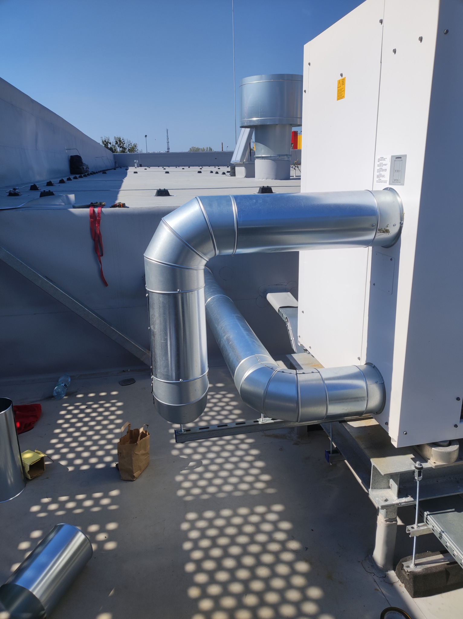 Instalacja wentylacyjna z rurami ze stali ocynkowanej, podłączona do białej jednostki klimatyzacyjnej na płaskim dachu, widoczny cień perforowanej konstrukcji.