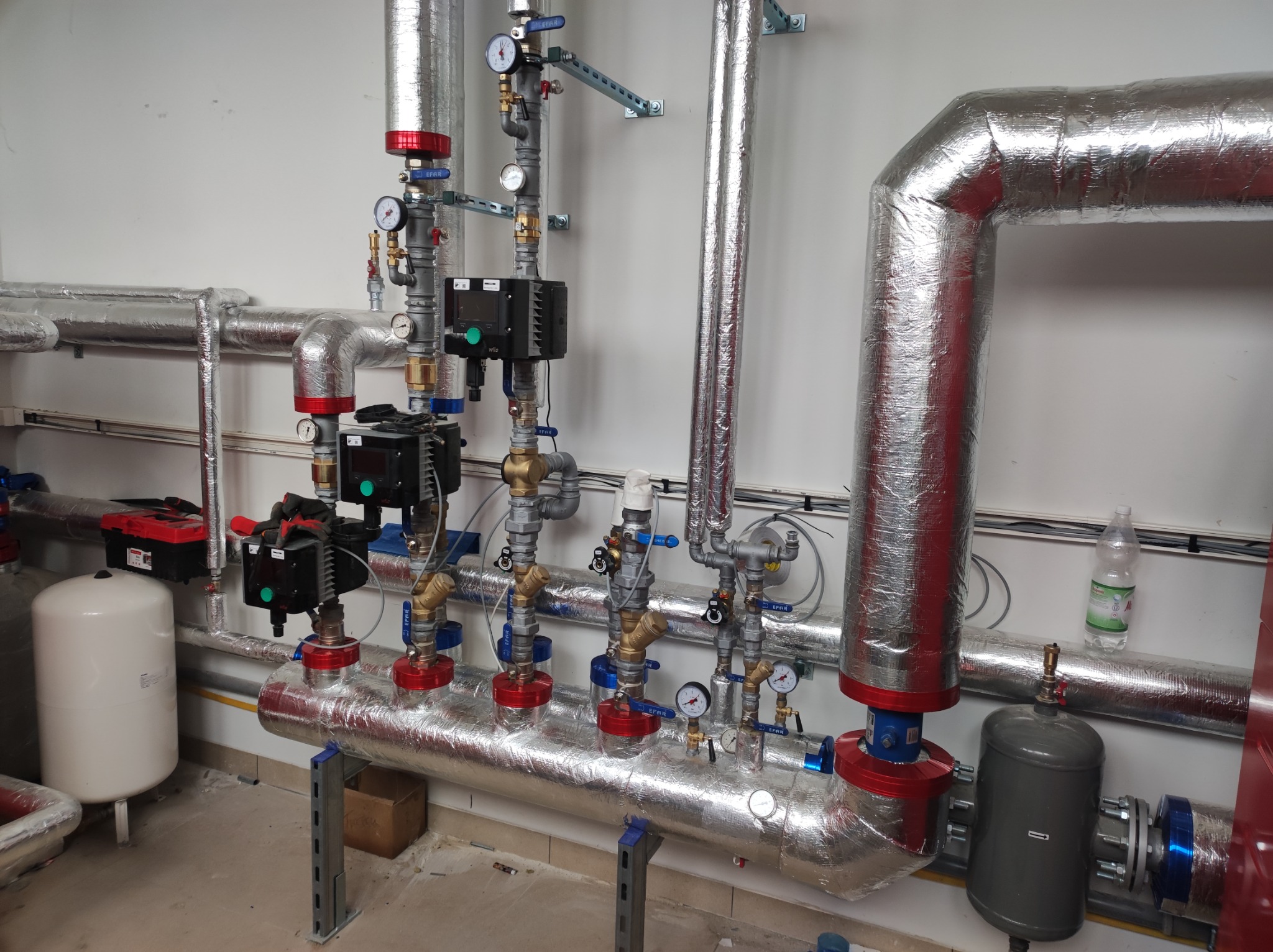 Kompleksowa instalacja grzewcza z rurami pokrytymi srebrną folią izolacyjną, zaworami EFAR z niebieskimi uchwytami i manometrami, zamontowana na białej ścianie w pomieszczeniu technicznym.