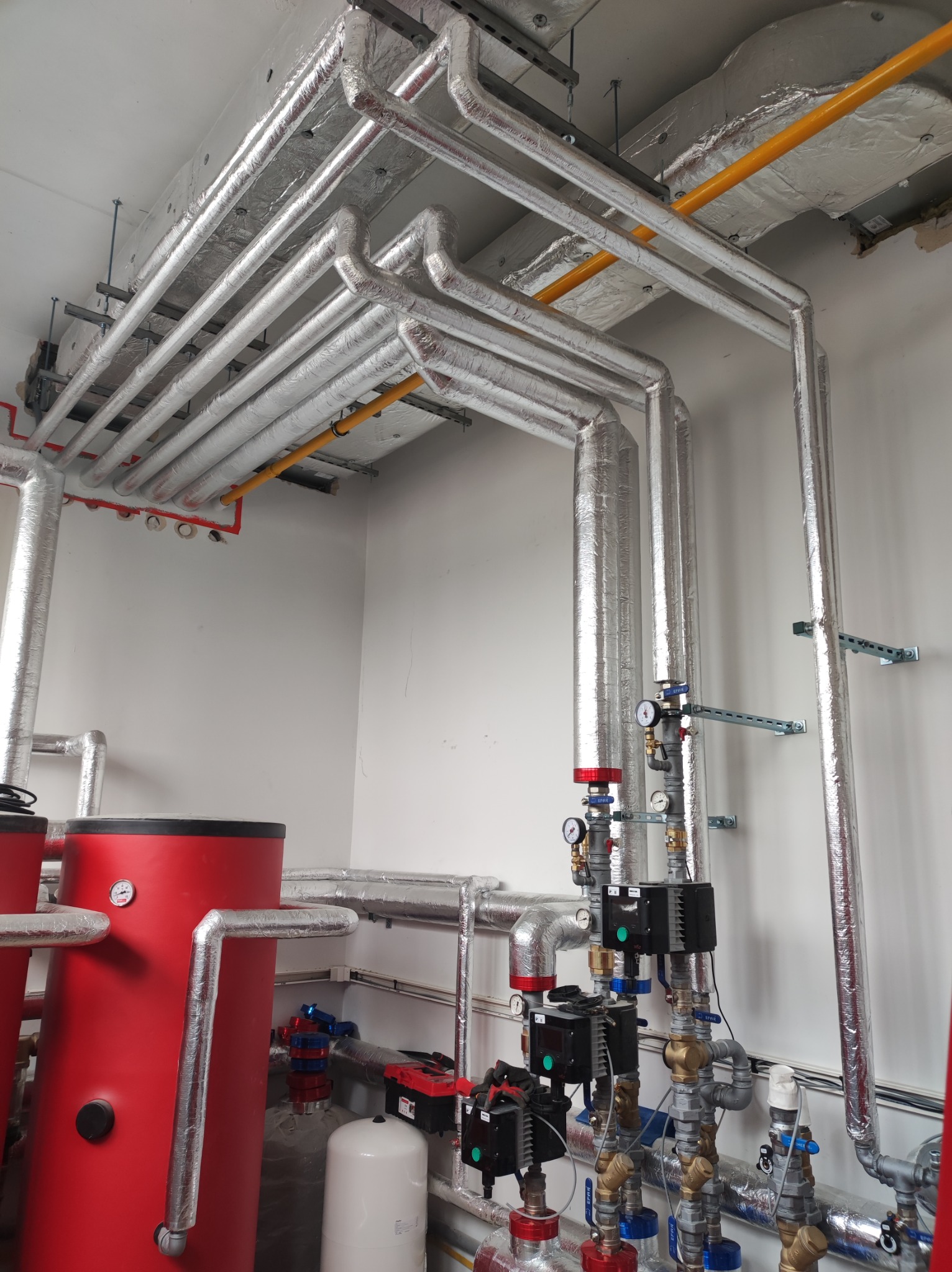 Instalacja centralnego ogrzewania z rurami w izolacji aluminiowej, czerwonymi zbiornikami i zaworami, widok na skomplikowany system rur pod sufitem.