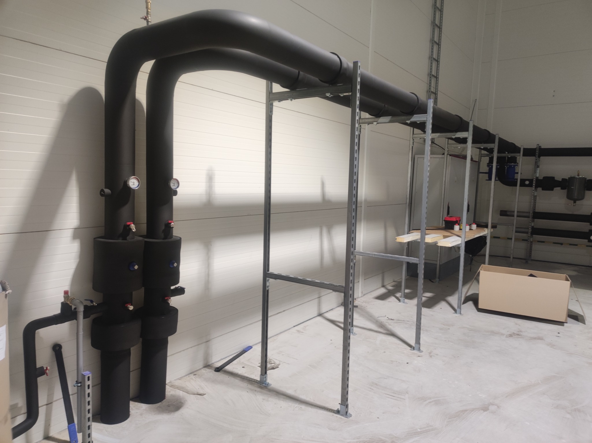 Czarne, zaizolowane rury grzewcze z manometrami, biegnące wzdłuż ściany z blachy trapezowej w hali, podtrzymywane przez metalowy stelaż.