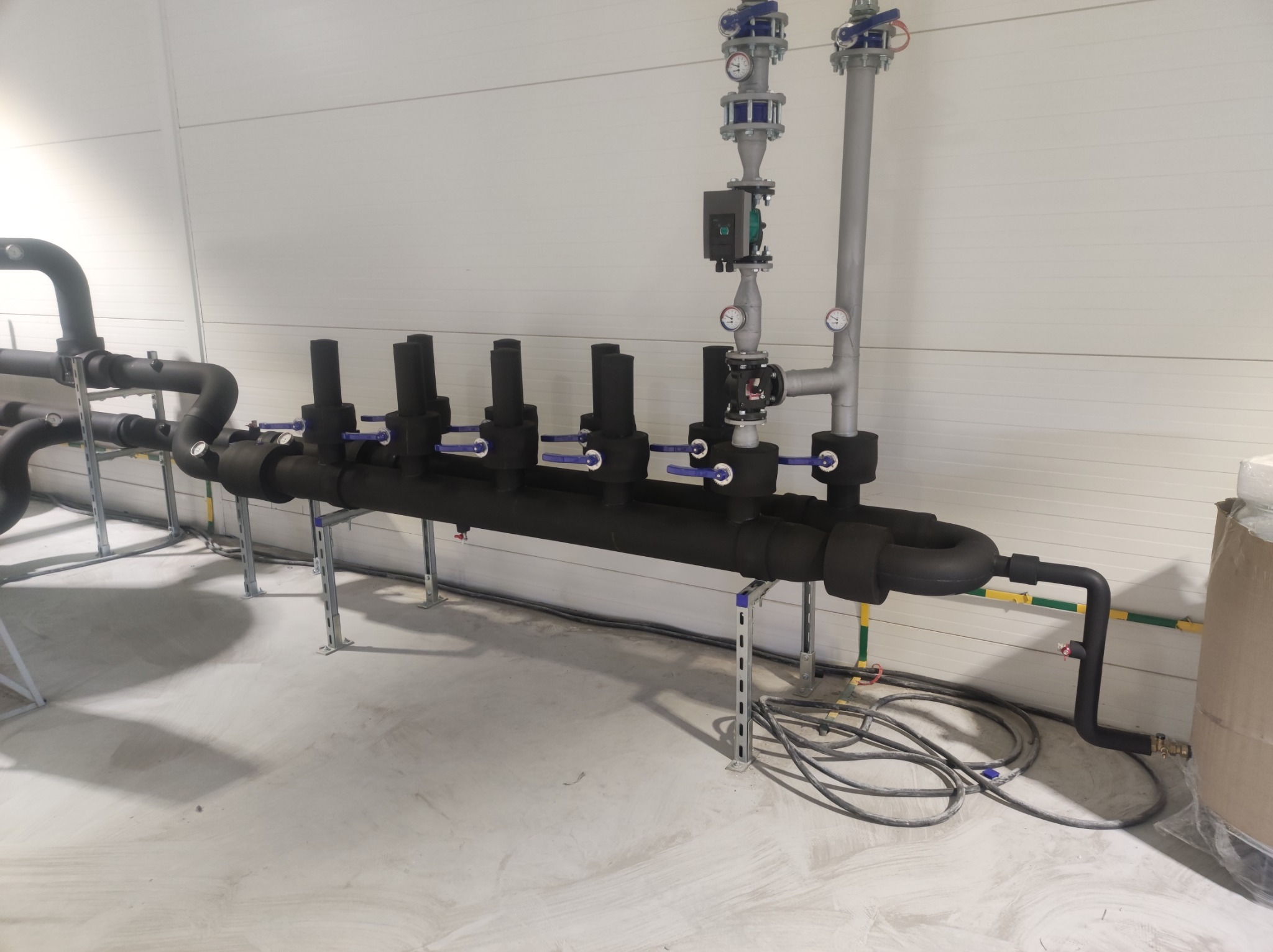 System rur z zaworami, izolowany czarną pianką, wsparty na metalowych wspornikach w jasnym pomieszczeniu przemysłowym z szarym betonowym podłożem.