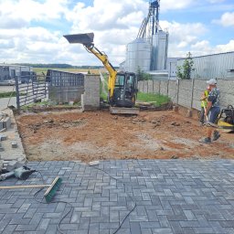 Roboty ziemne Paweł Pawluk - Żółta mini koparka na placu budowy, częściowo wyłożonym kostką brukową w jodełkę, w tle silosy i pracownicy z ubijarką.