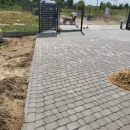 Ułożona szara kostka brukowa na podjeździe w Olszance, widoczna brama wjazdowa, piasek i pracownicy z taczkami w tle.