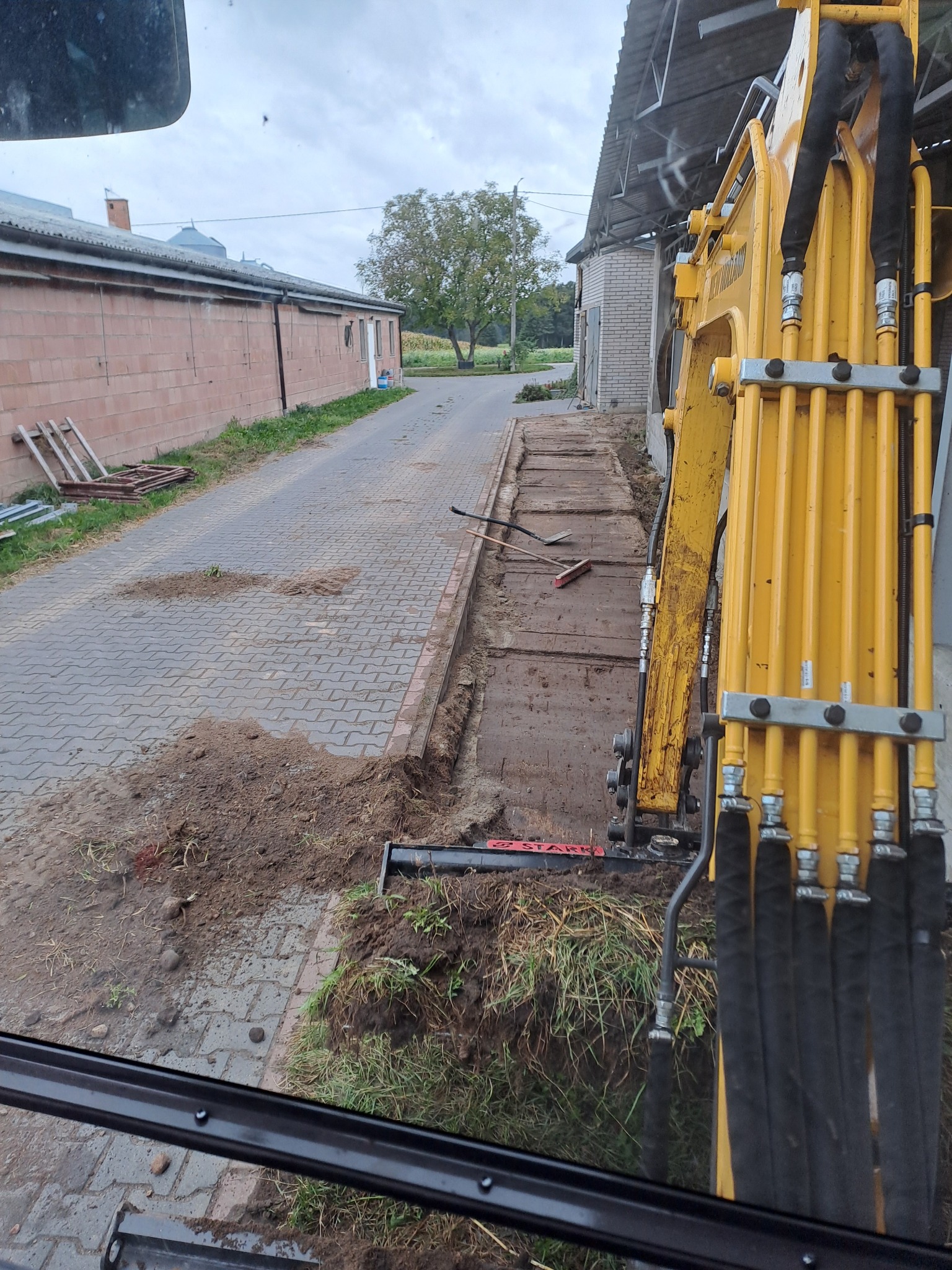 Żółta koparka na tle chodnika z kostki brukowej, przygotowuje teren pod instalacje, widok z kabiny operatora.