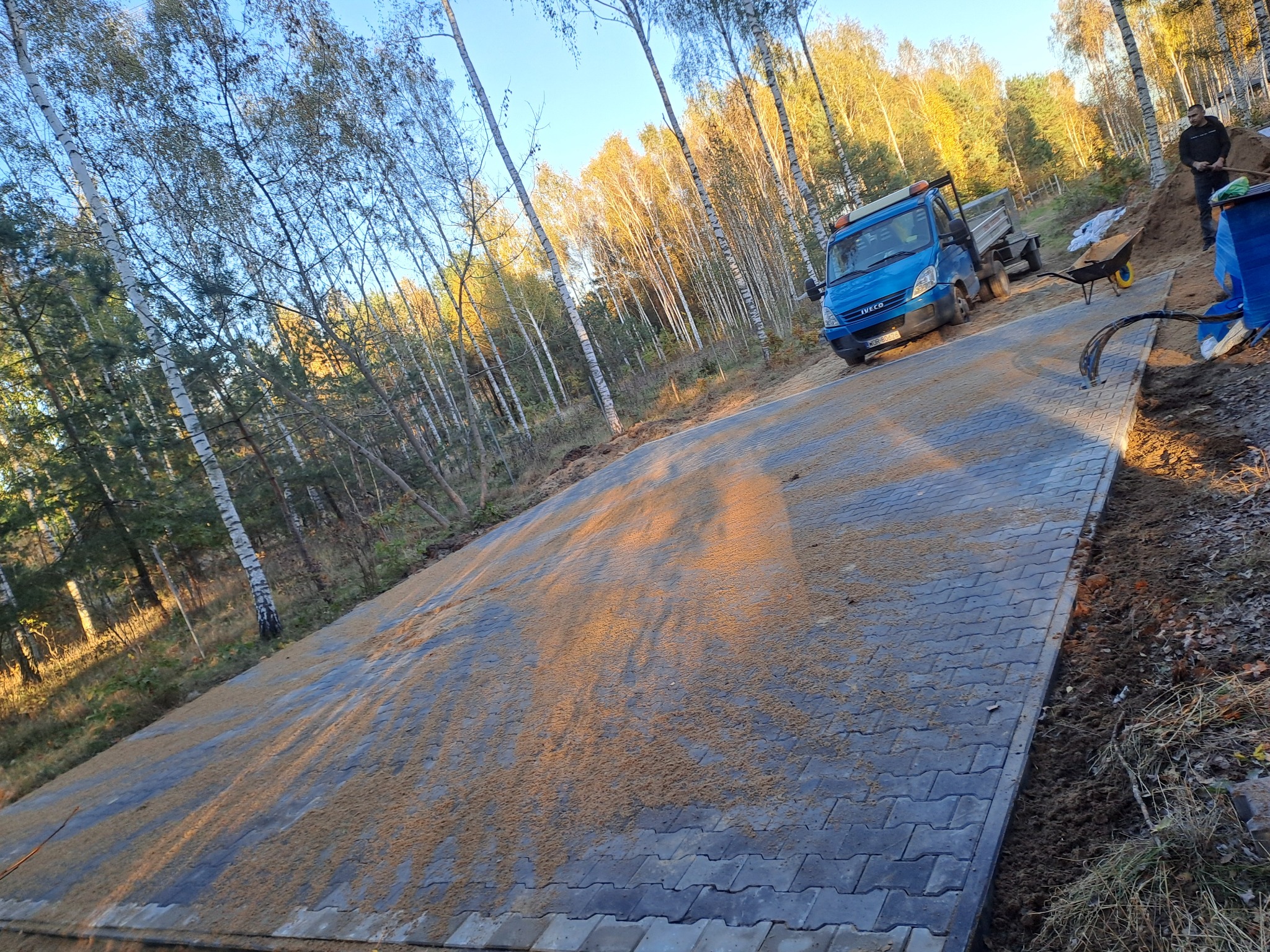Szeroki plac wyłożony szarą kostką brukową typu behaton, posypany piaskiem kwarcowym, z boku widoczny niebieski samochód dostawczy Iveco i taczka, w tle las brzozowy.