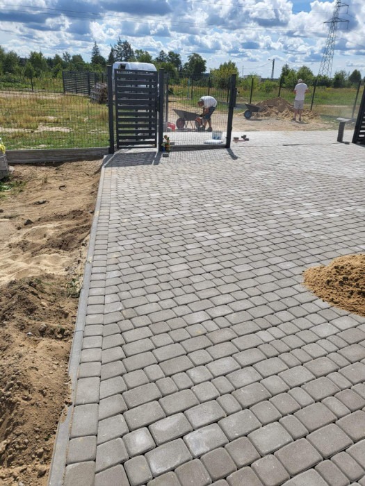 Ułożona szara kostka brukowa na podjeździe w Olszance, widoczna brama wjazdowa, piasek i pracownicy z taczkami w tle.