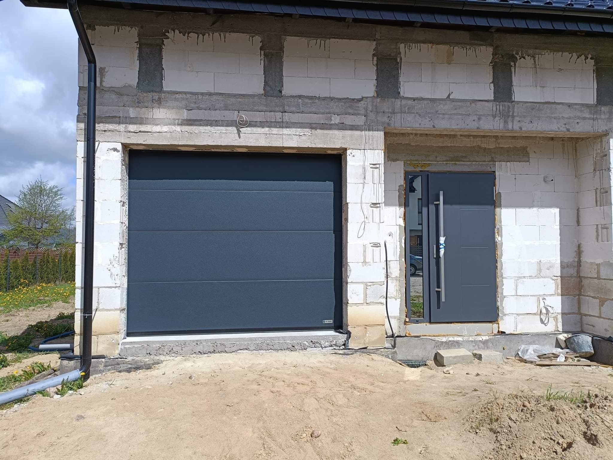 Nowa brama segmentowa w kolorze antracytowym zamontowana w surowym budynku z białego bloczka, widoczna również antracytowa stolarka drzwiowa, teren wokół w trakcie prac wykończeniowych.