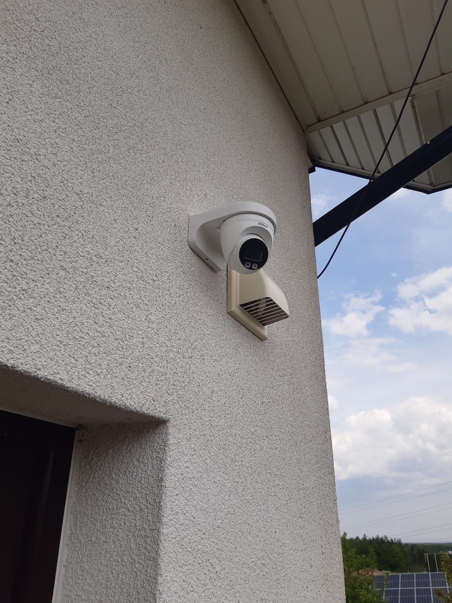 Kamera monitoringu IP zamontowana na teksturowanej, jasnej ścianie budynku, z widocznym fragmentem dachu i błękitnym niebem w tle. Dodatkowa osłona wentylacyjna pod kamerą. Widoczny fragment ramy...