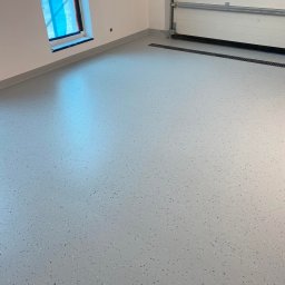 Perfect Coating System - Jasnoszara, gładka posadzka z drobnymi, ciemnymi plamkami w nowym pomieszczeniu z oknem zabezpieczonym folią i zamkniętą bramą garażową z kratką odpływową w podłodze.