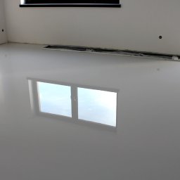 Perfect Coating System - Błyszcząca, gładka, jasnoszara posadzka w pustym pomieszczeniu z widocznymi oknami i ich odbiciem w powierzchni podłogi, zabezpieczonymi elementami przy ścianie.