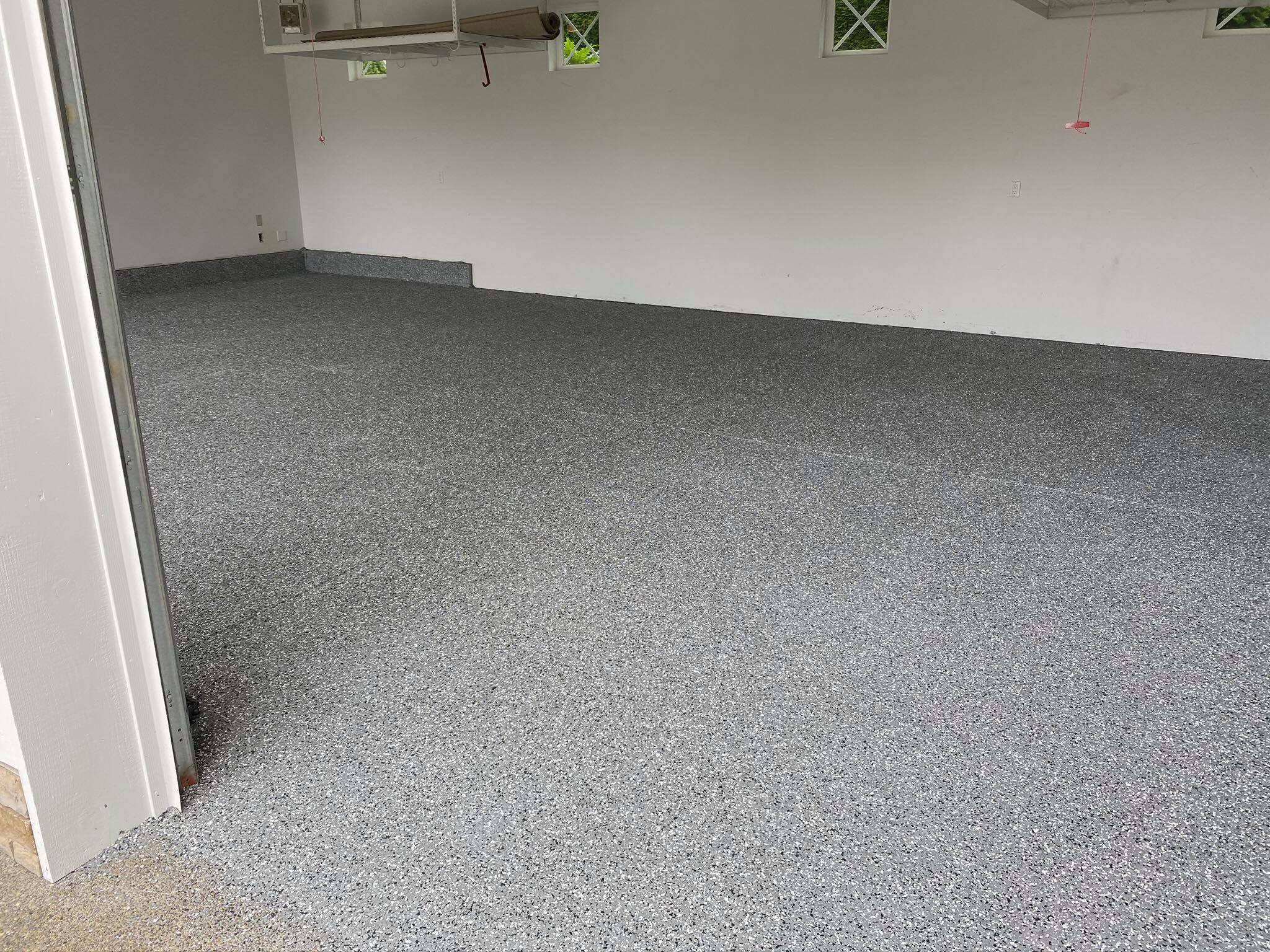 Wykończona posadzka żywiczna w garażu o szarym, nakrapianym wzorze, widoczna z perspektywy wejścia, z białymi ścianami i oknami w górnej części ściany.