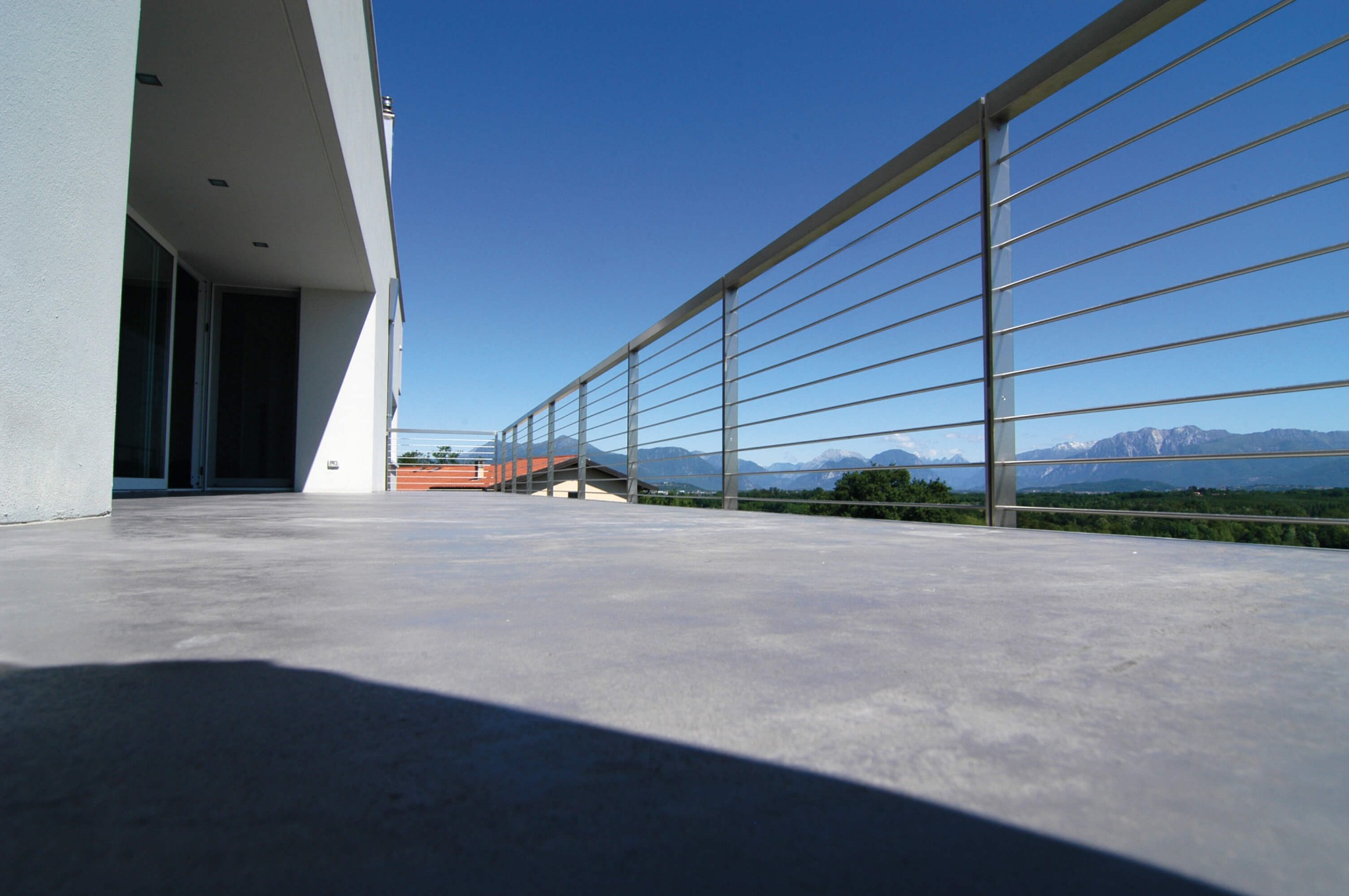 Szara, gładka posadzka balkonu z metalową balustradą na tle błękitnego nieba i górskiego krajobrazu.