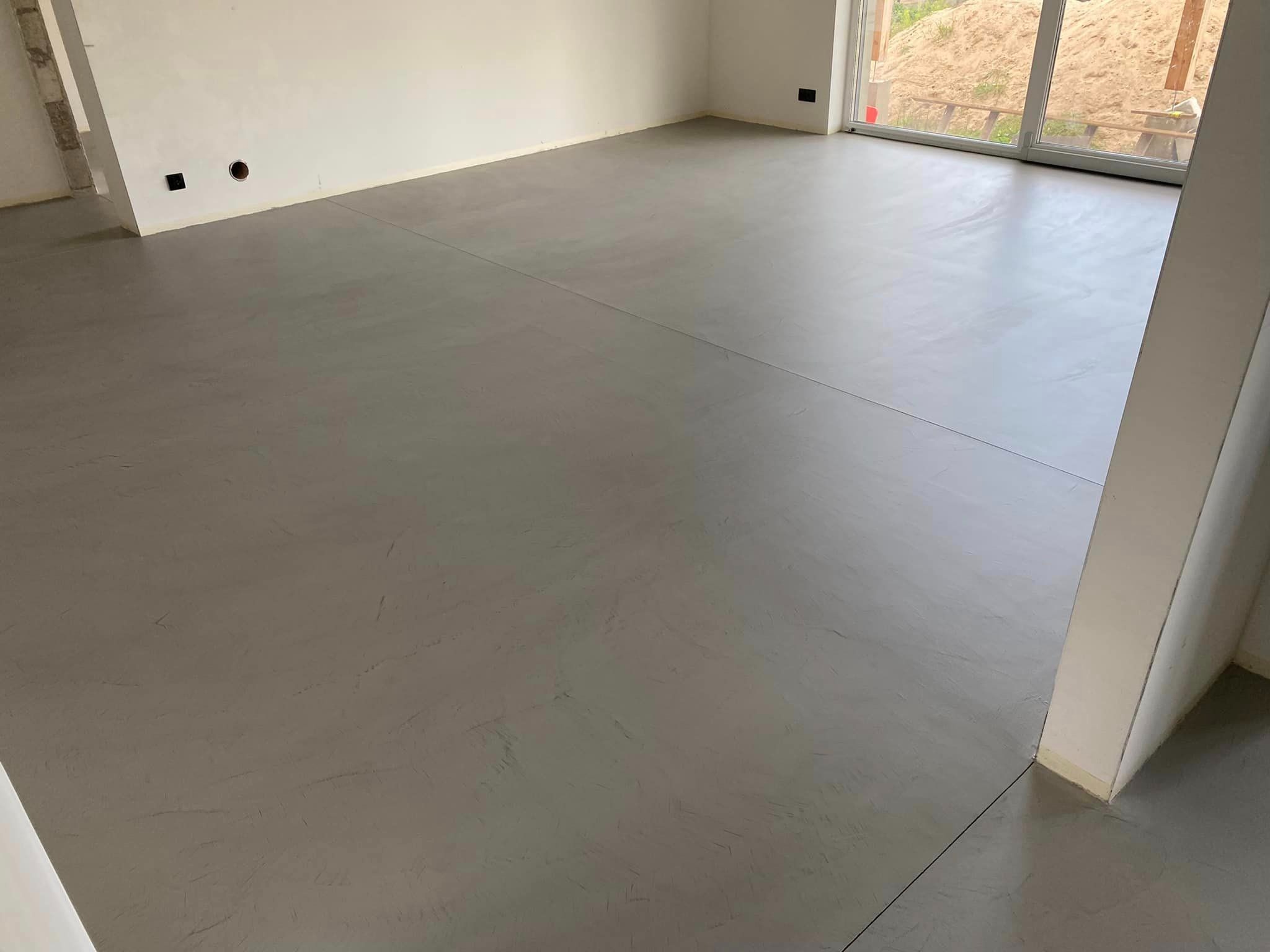 Szara, gładka posadzka w nowym domu w Kunowie, z widocznymi dylatacjami, przez okno widać nasyp ziemi.