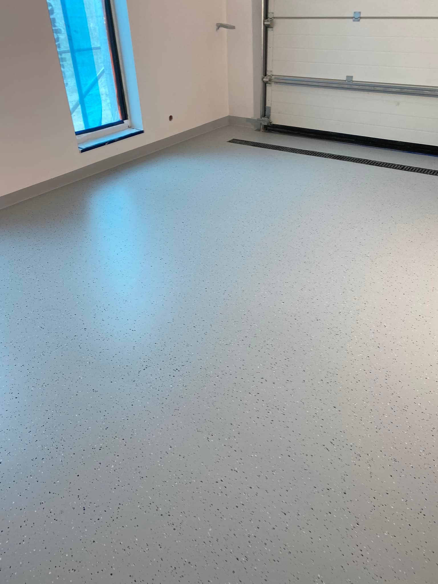 Jasnoszara, gładka posadzka z drobnymi, ciemnymi plamkami w nowym pomieszczeniu z oknem zabezpieczonym folią i zamkniętą bramą garażową z kratką odpływową w podłodze.