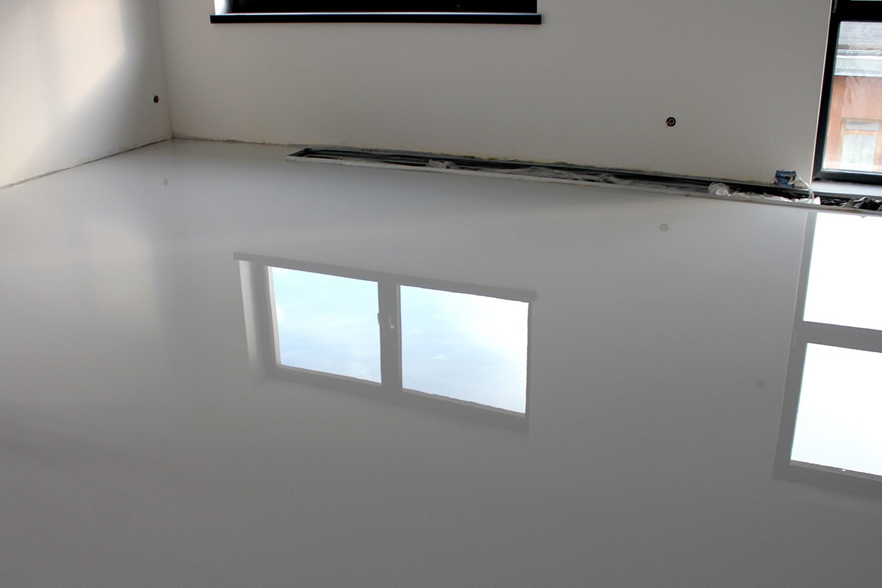 Błyszcząca, gładka, jasnoszara posadzka w pustym pomieszczeniu z widocznymi oknami i ich odbiciem w powierzchni podłogi, zabezpieczonymi elementami przy ścianie.