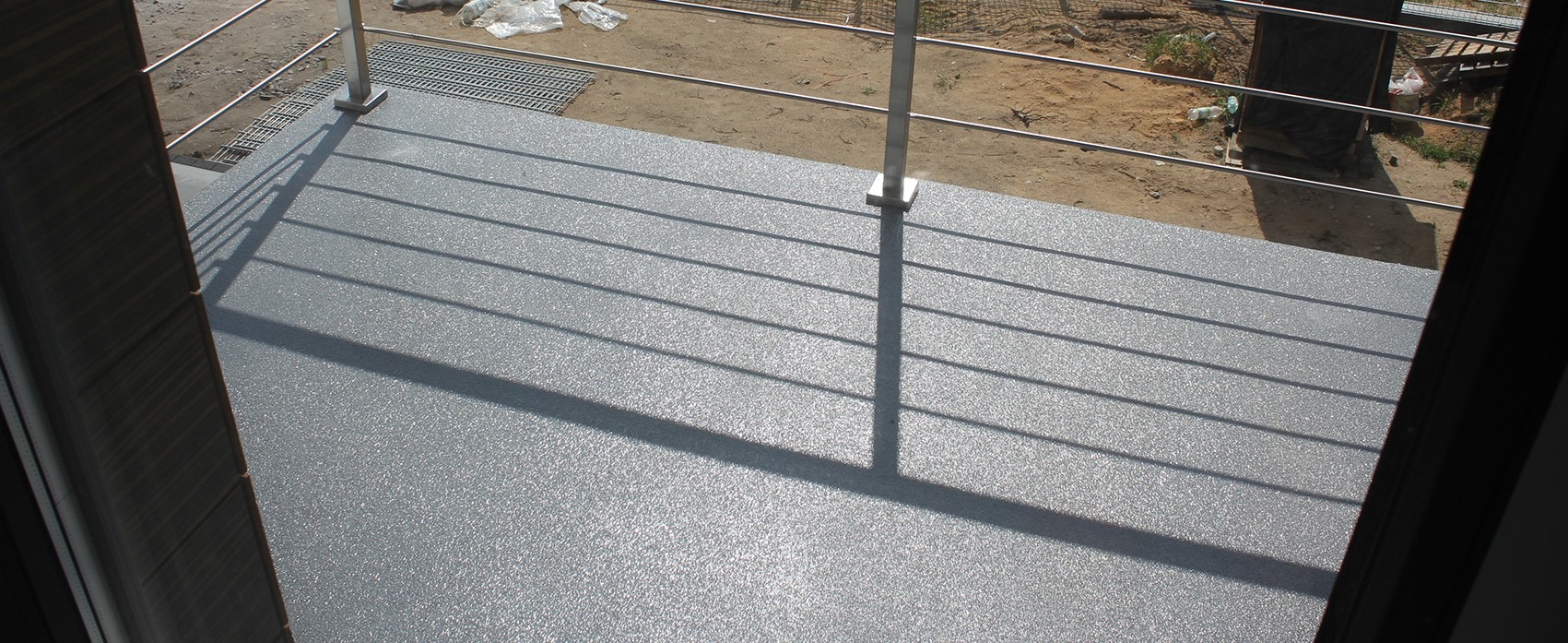 Szara posadzka żywiczna na balkonie, widoczne cienie barierki i fragment otoczenia zewnętrznego z ziemią i workami.