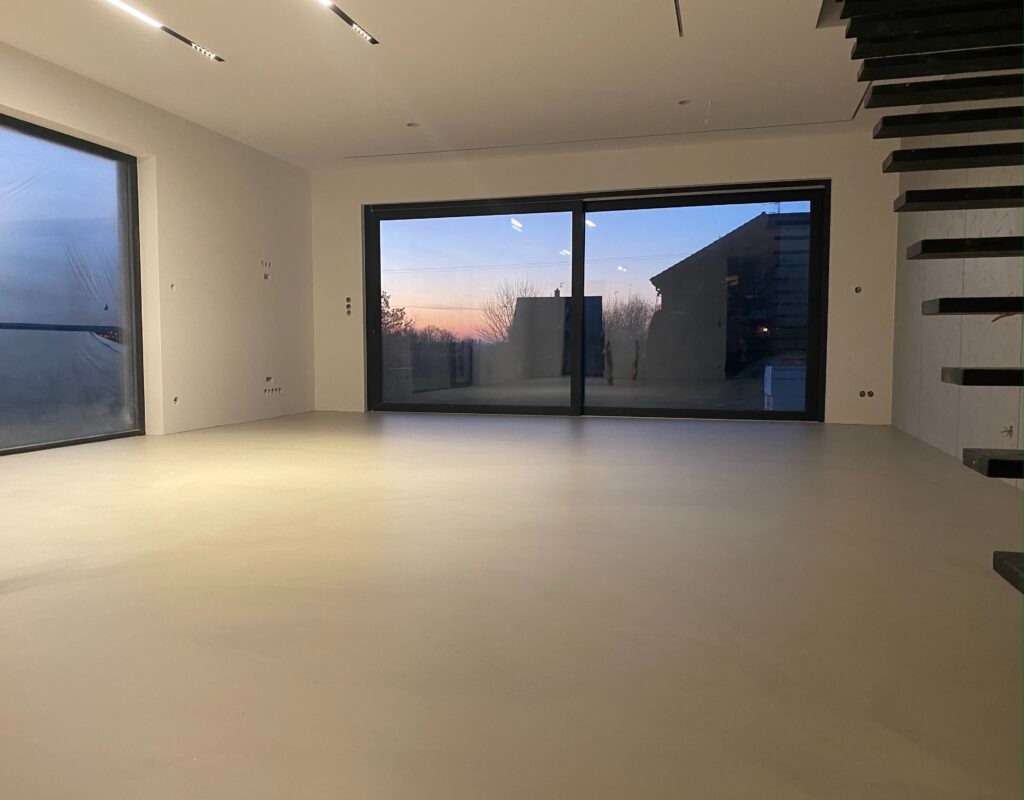 Jasnoszara, gładka posadzka w przestronnym wnętrzu z dużymi oknami, widoczne schody bez poręczy, zachód słońca w tle.