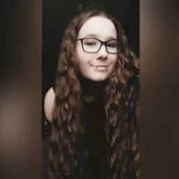 Portret uśmiechniętej kobiety w okularach i z piercingiem w ustach, z długimi, kręconymi brązowymi włosami na ciemnym tle.