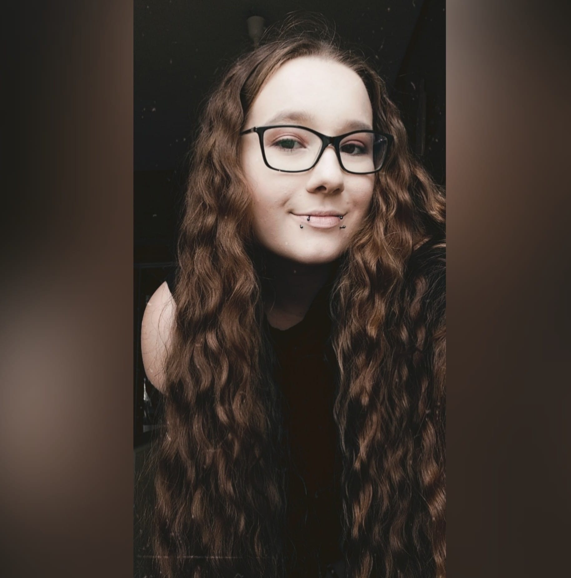 Portret uśmiechniętej kobiety w okularach i z piercingiem w ustach, z długimi, kręconymi brązowymi włosami na ciemnym tle.