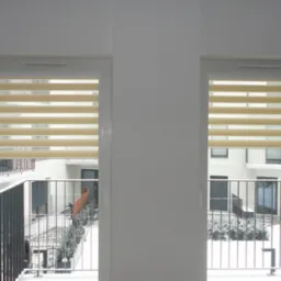 Dwa okna PCV z zamontowanymi beżowymi roletami typu dzień i noc, widok z wnętrza na balkon i dziedziniec osiedla w zimowej scenerii.