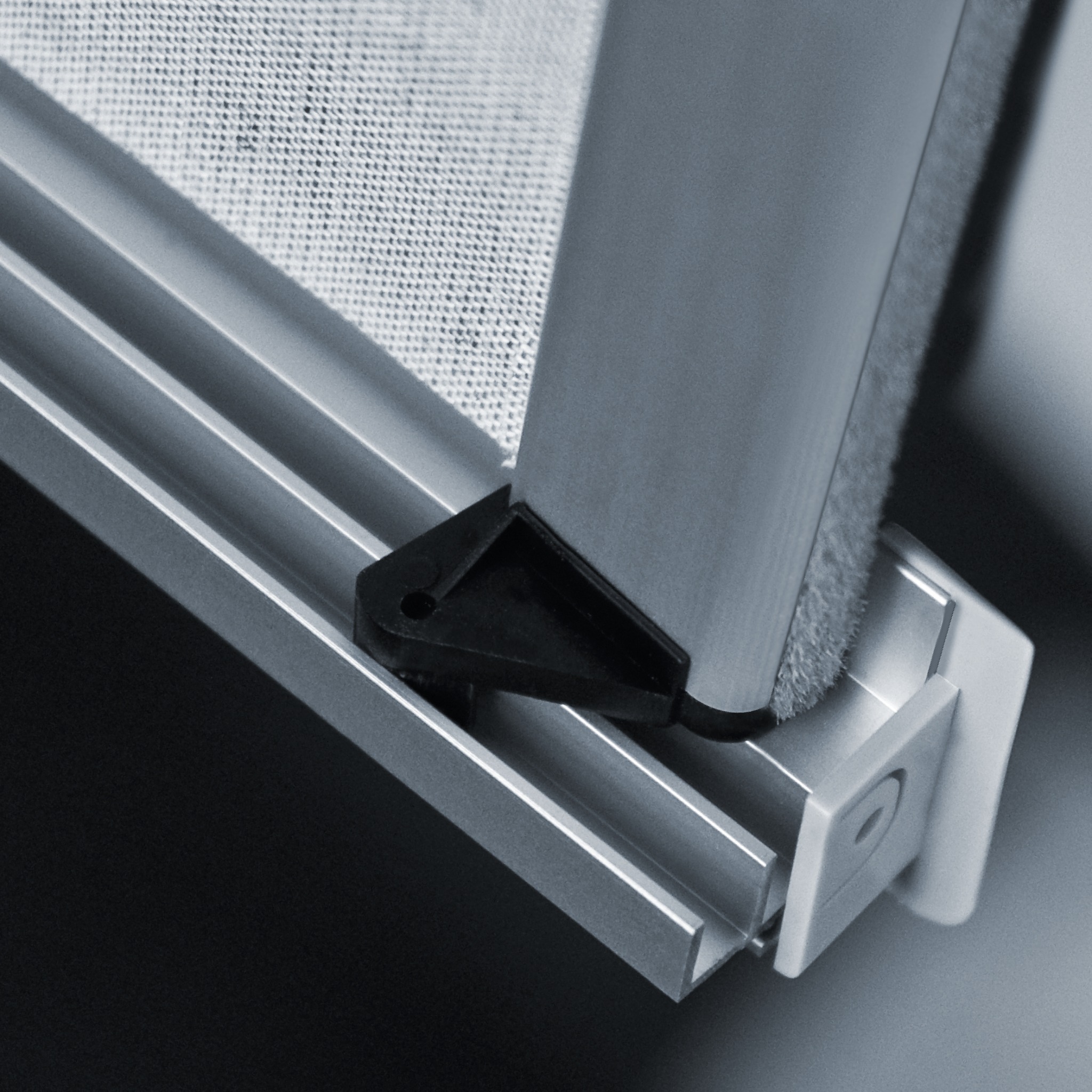 Szczegółowe zbliżenie na mechanizm prowadzący tekstylną osłonę okienną z aluminiową ramą i czarnym elementem mocującym.