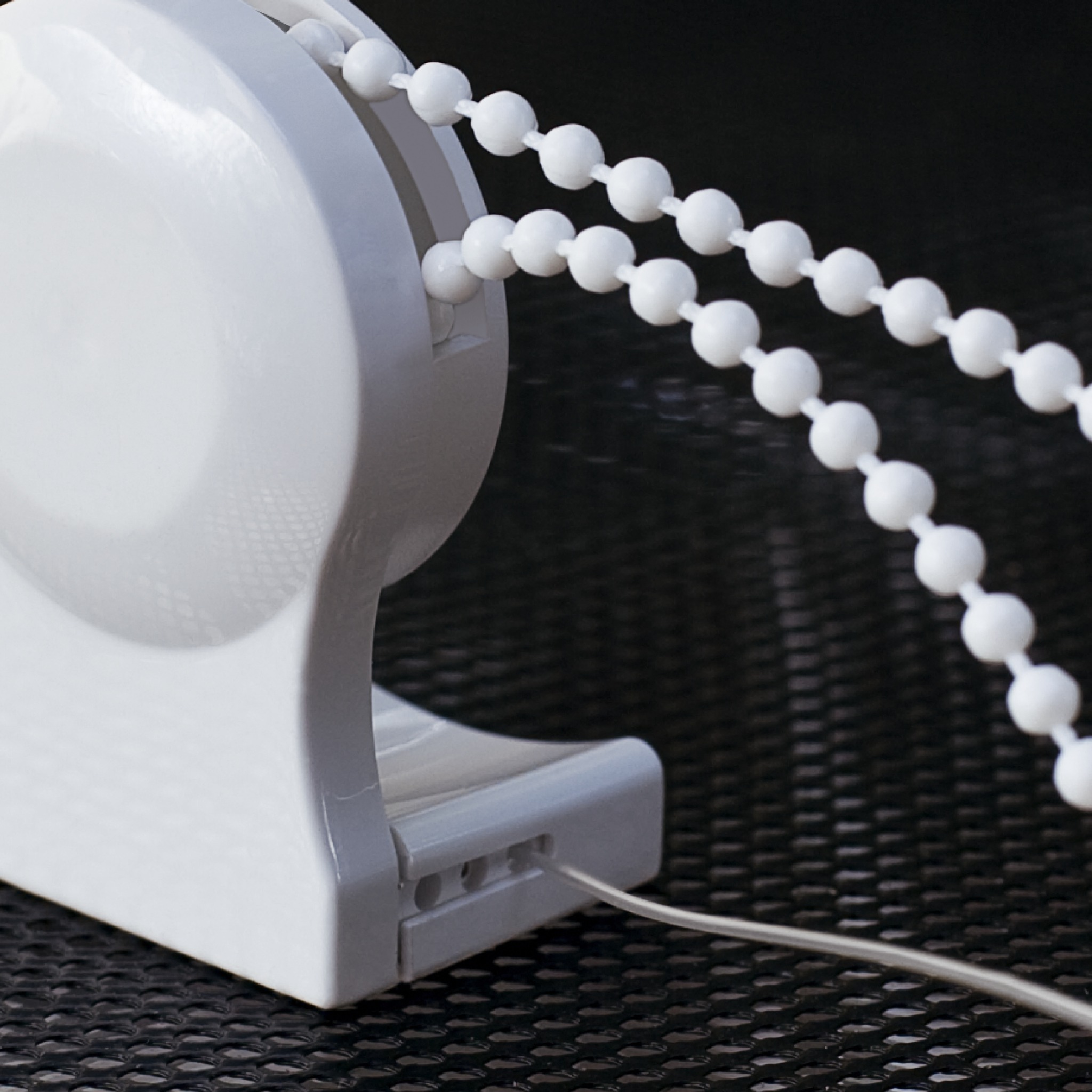 Mechanizm sterujący z białym łańcuszkiem kulkowym do podnoszenia i opuszczania osłony okiennej, widoczny przewód na tle ciemnej, teksturowanej powierzchni.