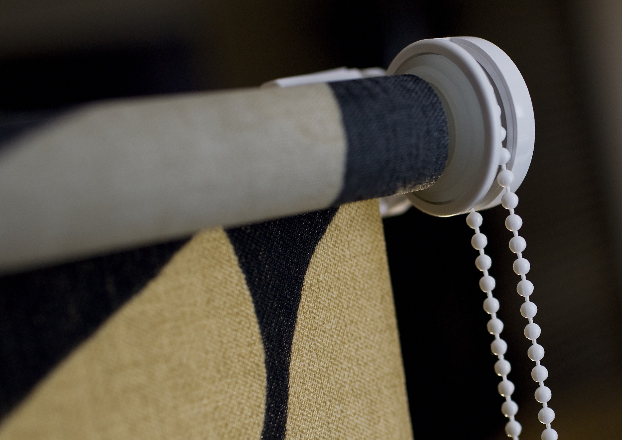 Fragment zwiniętej rolety z geometrycznym wzorem w odcieniach beżu i czerni, widoczny mechanizm z białym łańcuszkiem kulkowym.