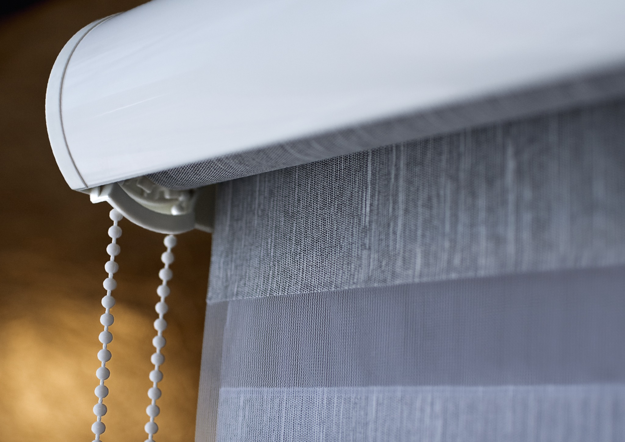 Detale tekstylnej rolety dzień noc z białym mechanizmem i łańcuszkiem kulkowym na tle ściany w ciepłym odcieniu.