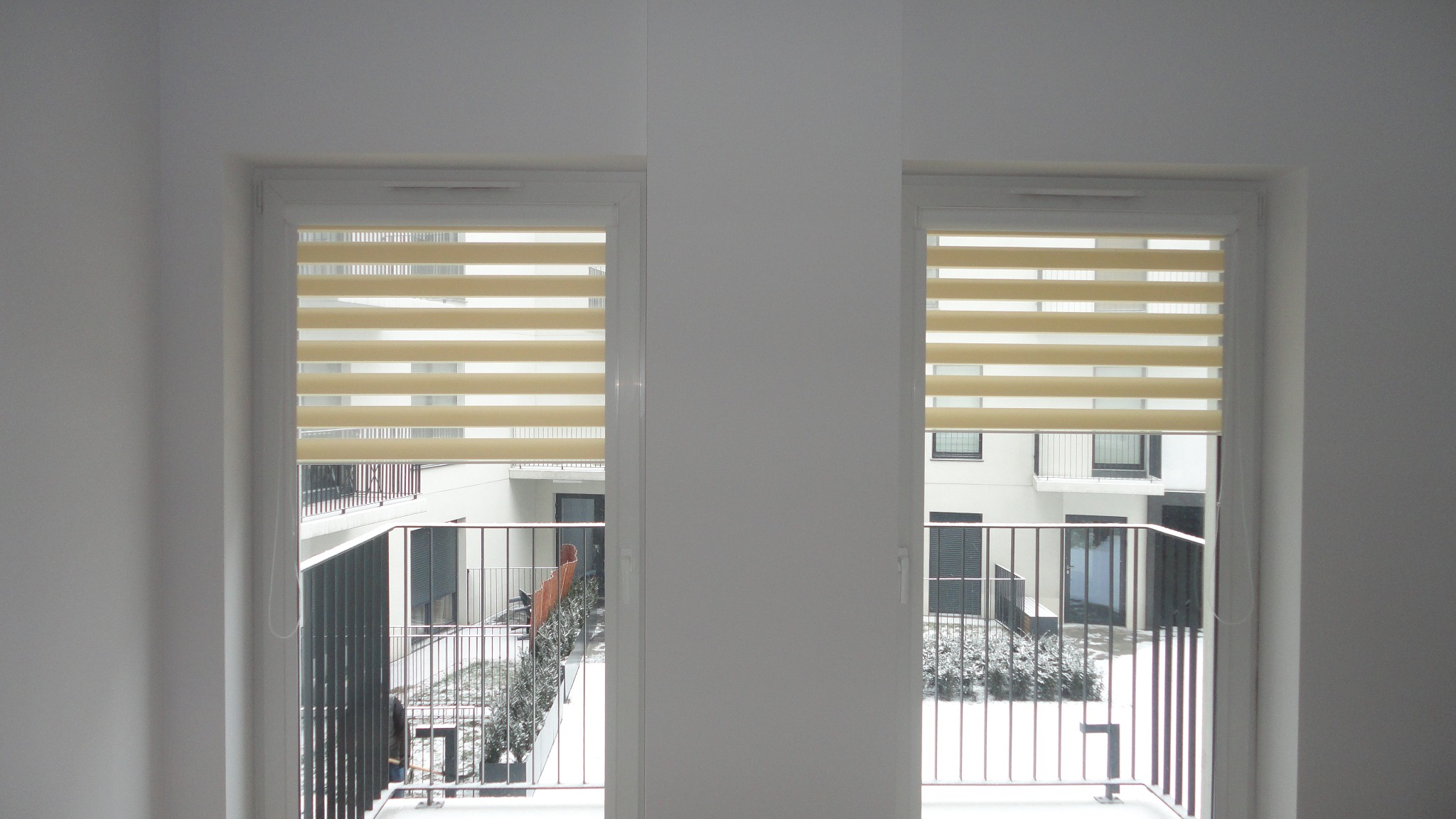 Dwa okna PCV z zamontowanymi beżowymi roletami typu dzień i noc, widok z wnętrza na balkon i dziedziniec osiedla w zimowej scenerii.