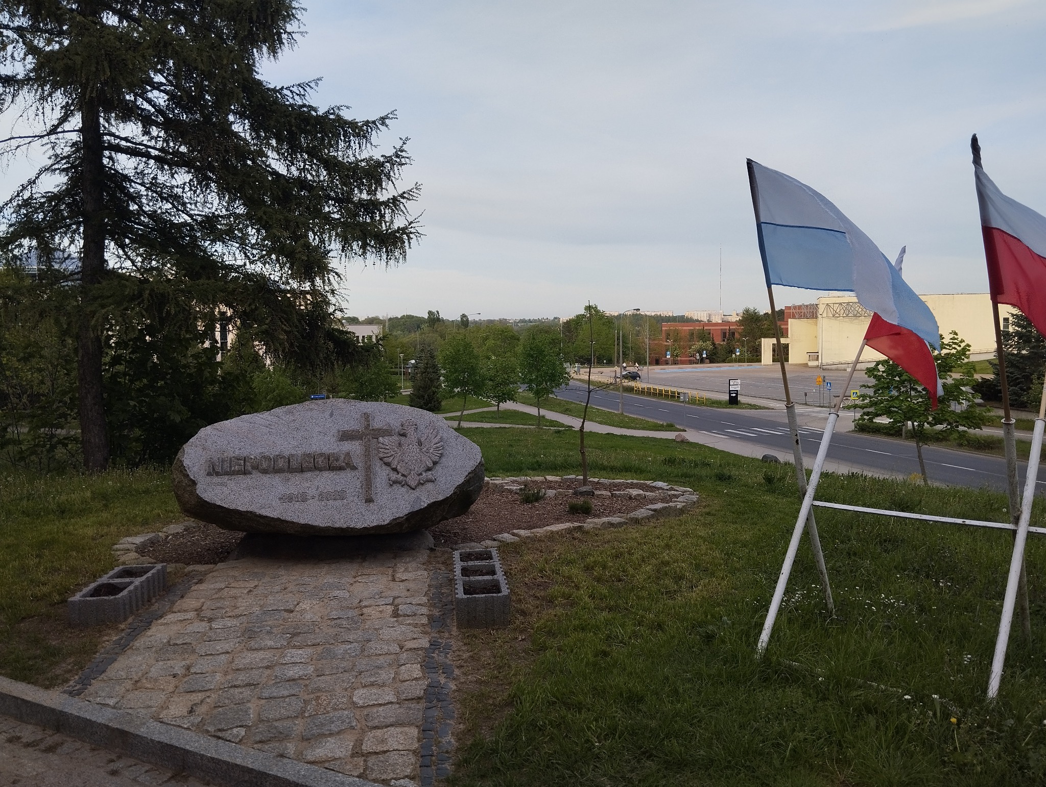 Kamień z napisem 'Niepodległa 1918-2022' i symbolem orła, otoczony zielenią i flagami, na tle miejskiego krajobrazu.