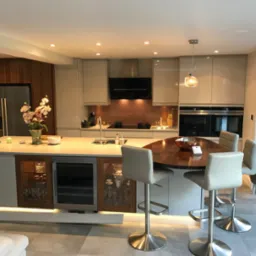 Nowoczesna kuchnia z wyspą, barkiem z drewnianym blatem, szarymi szafkami i oświetleniem LED pod szafkami. Widoczna zabudowa ze stali nierdzewnej, wbudowane sprzęty AGD i okno z widokiem na ogród.