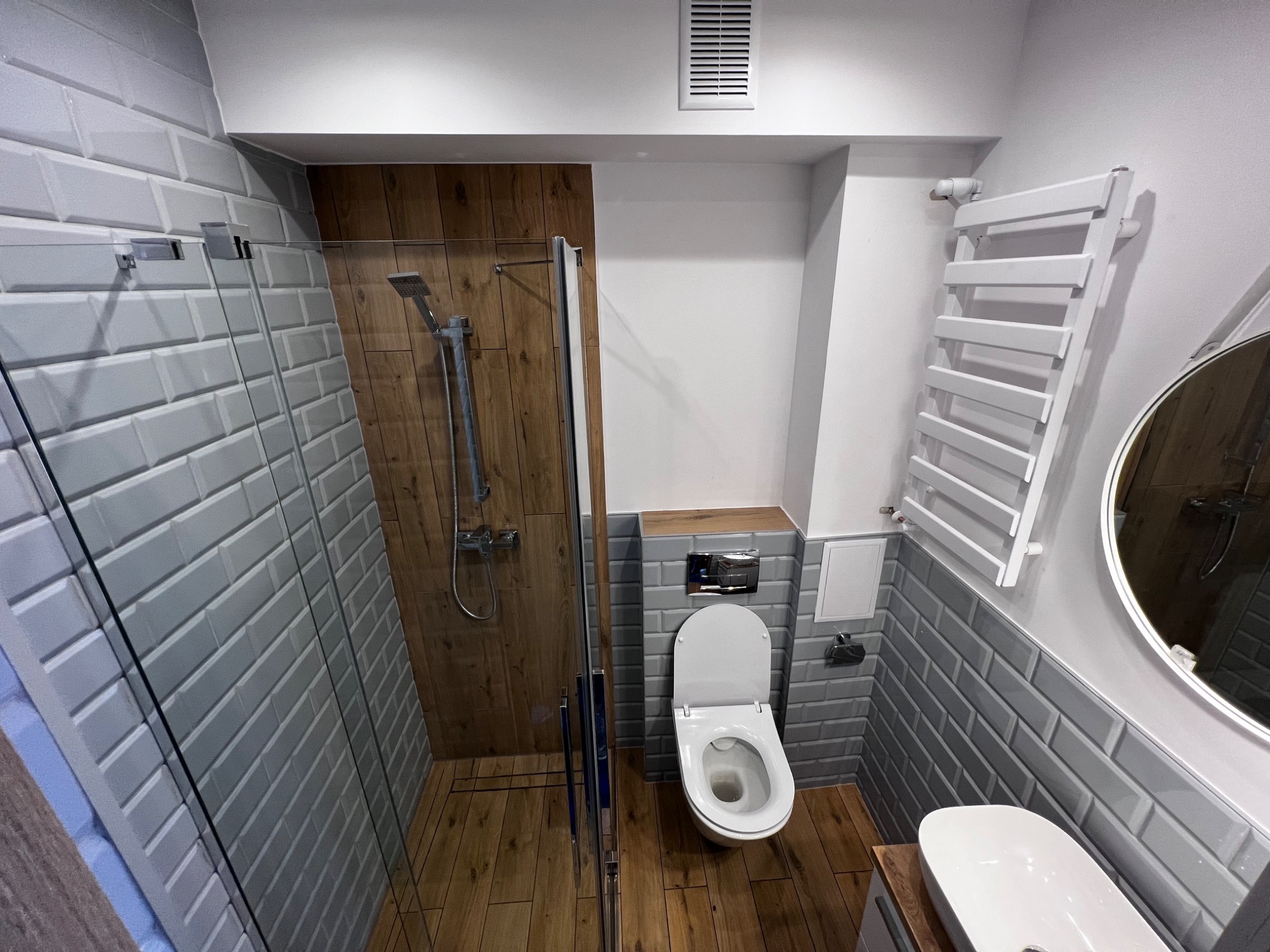 Nowoczesna łazienka po remoncie z szarymi, cegiełkowymi płytkami, kabiną prysznicową z drewnianym panelem, podwieszaną toaletą i grzejnikiem drabinkowym.