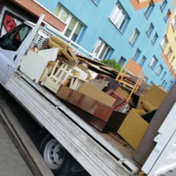 Biała skrzyniowa ciężarówka wypełniona meblami i odpadami drewnianymi na tle kolorowego bloku mieszkalnego z graffiti na kabinie.