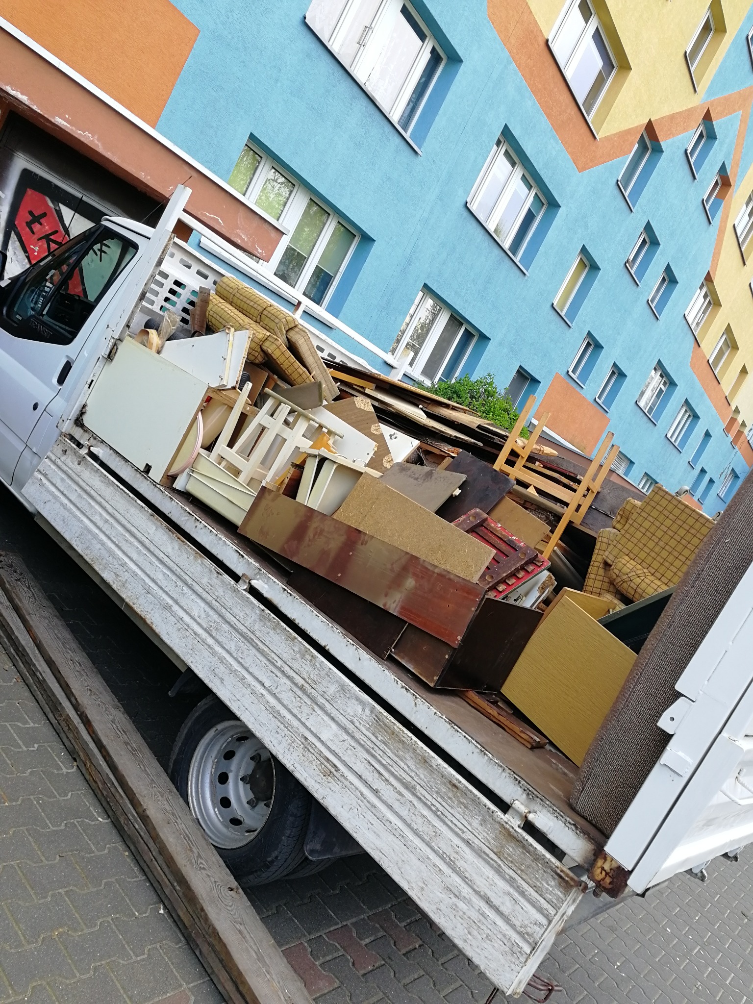 Biała skrzyniowa ciężarówka wypełniona meblami i odpadami drewnianymi na tle kolorowego bloku mieszkalnego z graffiti na kabinie.