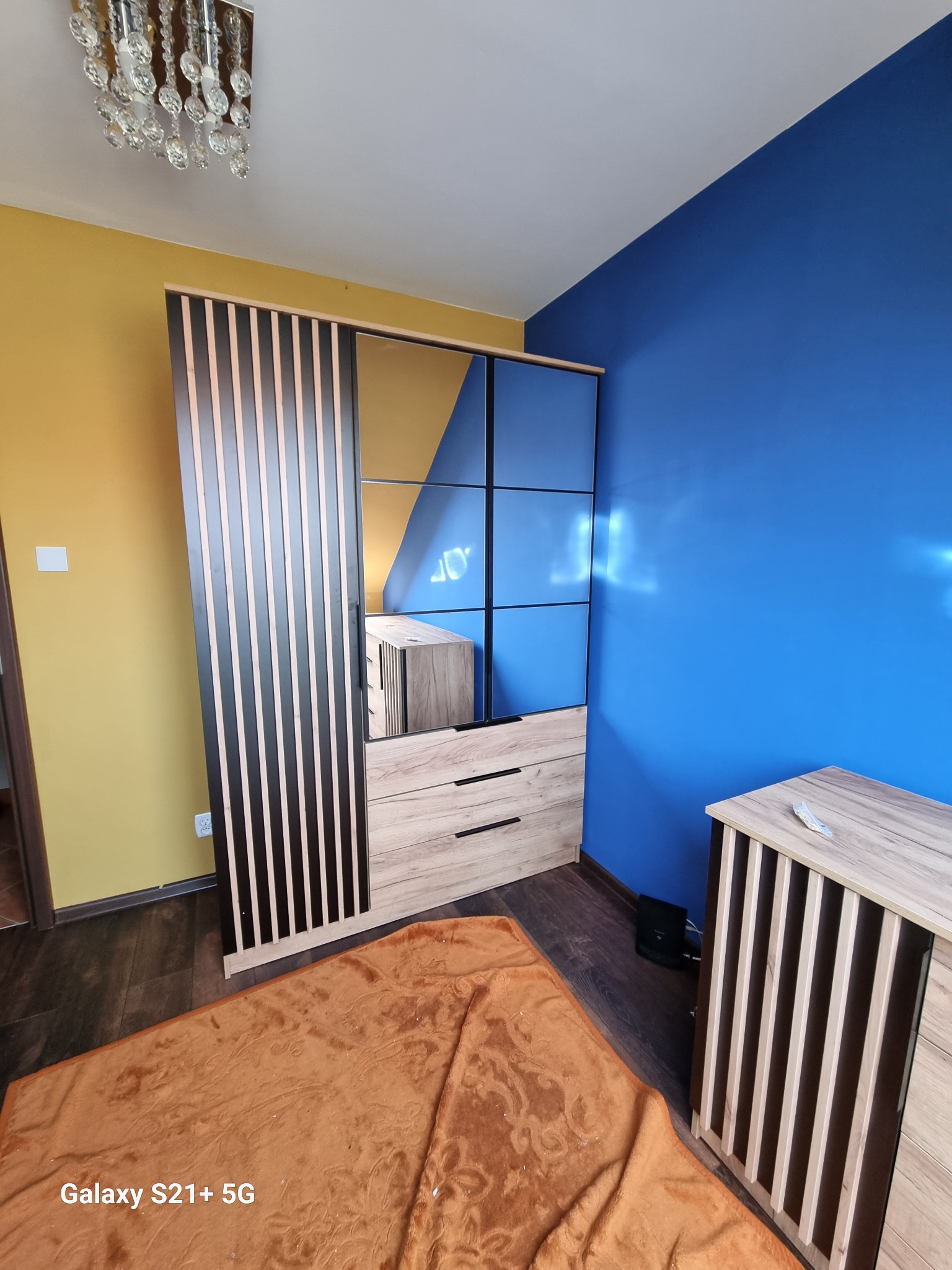 Nowoczesna szafa z lustrem i drewnianymi lamelami w pokoju z żółtą i niebieską ścianą, widoczny fragment brązowego dywanu.