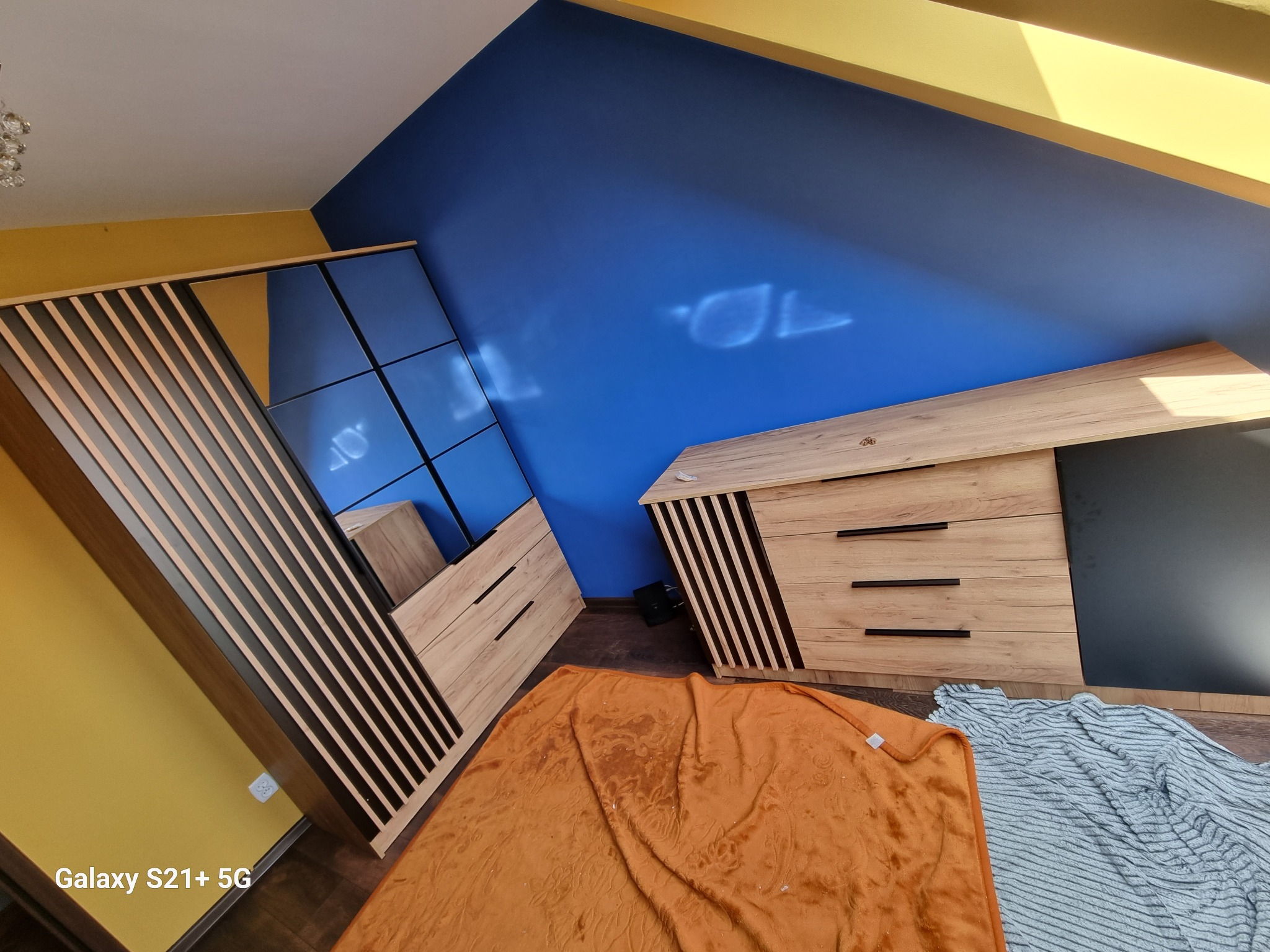 Szafa przesuwna z lustrem i drewnianymi lamelami, komoda z szufladami w pokoju z niebiesko-żółtymi ścianami na poddaszu.