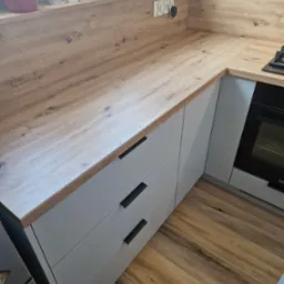 Fragment nowoczesnej kuchni z drewnianym blatem, szarymi szafkami z czarnymi uchwytami i piekarnikiem w zabudowie. Na blacie widoczne gniazdka elektryczne i płyta gazowa. Podłoga wyłożona panelami...