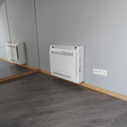 SWOKI Nina brusenska - Biały klimatyzator ścienny zamontowany w rogu pokoju z szarą ścianą i ciemną podłogą, widoczny również w lustrzanym odbiciu.