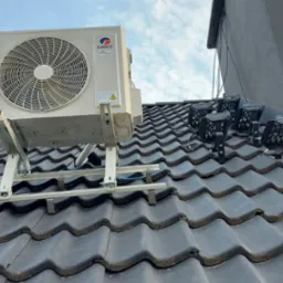 Biała jednostka zewnętrzna klimatyzatora marki Gree zamontowana na metalowym stelażu na ciemnoszarym dachu z dachówki ceramicznej, z widocznymi elementami instalacji fotowoltaicznej w tle.