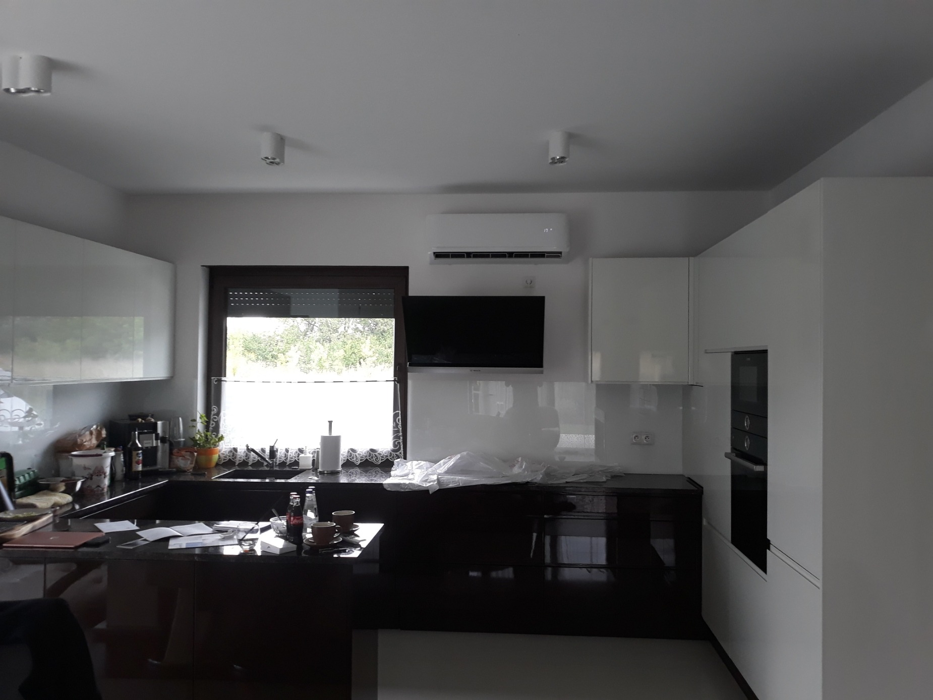Nowoczesna kuchnia z białymi szafkami i czarnym blatem, widoczny klimatyzator ścienny nad telewizorem.