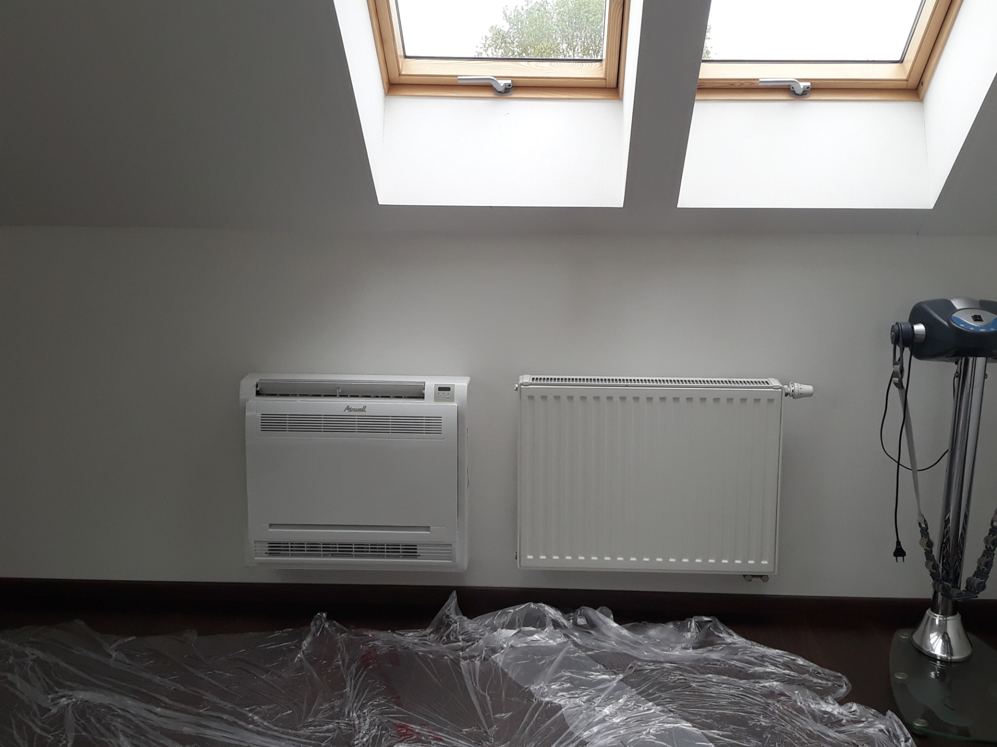 Biały klimatyzator podłogowy marki Airwell i grzejnik panelowy zamontowane na ścianie w pokoju, podłoga zabezpieczona folią malarską, widoczne okna dachowe.