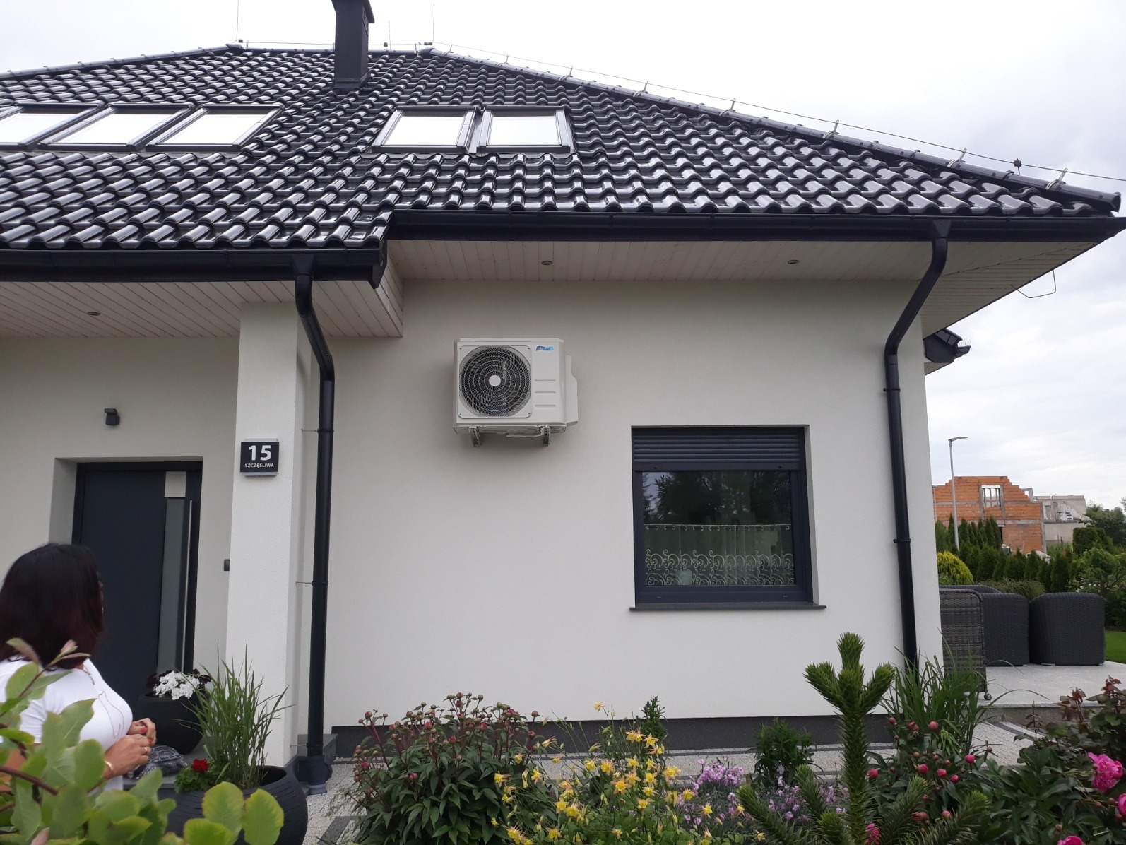 Biała elewacja domu jednorodzinnego z zamontowaną jednostką zewnętrzną klimatyzacji, widok z poziomu ogrodu z kwitnącymi kwiatami.